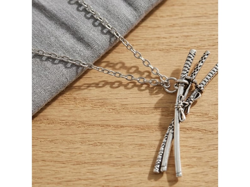May include: Silver-toned necklace featuring a pendant of three miniature katana swords. The swords are intricately detailed and tied together with a decorative element. The chain is a simple link style, resting on a gray fabric and wooden surface.