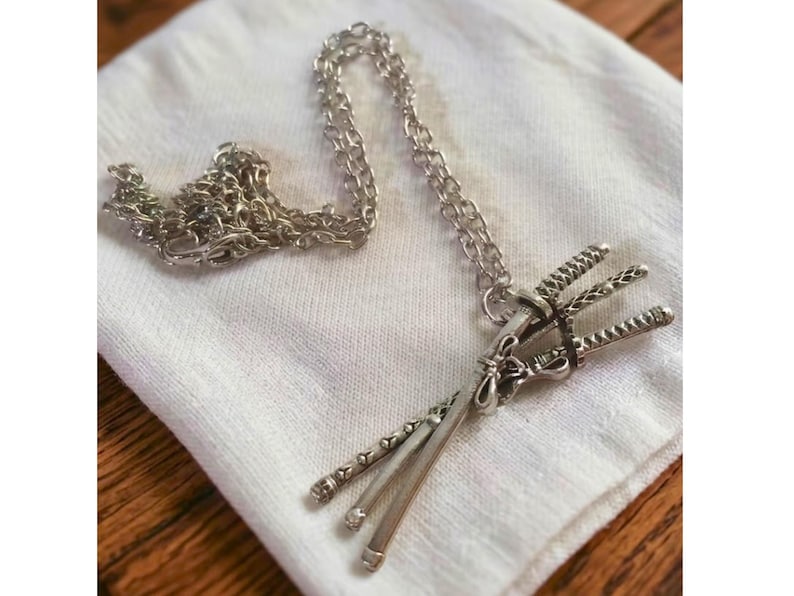 May include: A silver-colored pendant necklace featuring a stylized design of three miniature swords bound together. The pendant hangs from a delicate chain, resting on a white cloth. The necklace is a unique accessory.