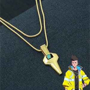 May include: A gold-coloured necklace featuring a double-layered snake chain and a geometric pendant with a small turquoise accent. The pendant has a smooth, angular shape. An illustration of a character wearing a yellow jacket is positioned below.