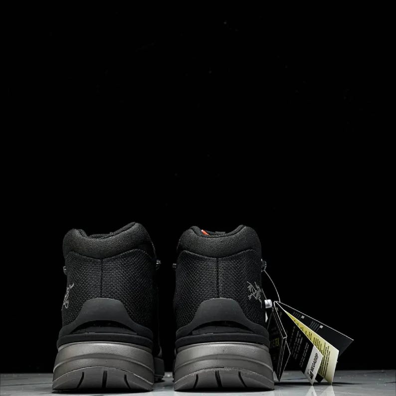 May include: Two black hiking boots with grey soles, seen from behind. The boots feature a textured upper and a small logo. A tag is attached to one boot.