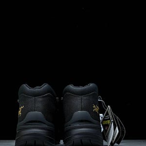 May include: Pair of black hiking boots with a textured upper and thick soles. The boots have a gold logo on the side and a tag that reads "GORE-TEX". The boots are designed for outdoor activities.
