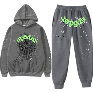Pu&ograve; includere: Un completo composto da felpa con cappuccio e pantaloni della tuta grigi. La felpa con cappuccio presenta una grafica a ragnatela e la scritta "sp5der" in verde neon. Anche i pantaloni della tuta hanno la scritta "sp5der" e dettagli a stella. Il completo &egrave; realizzato in materiale morbido.