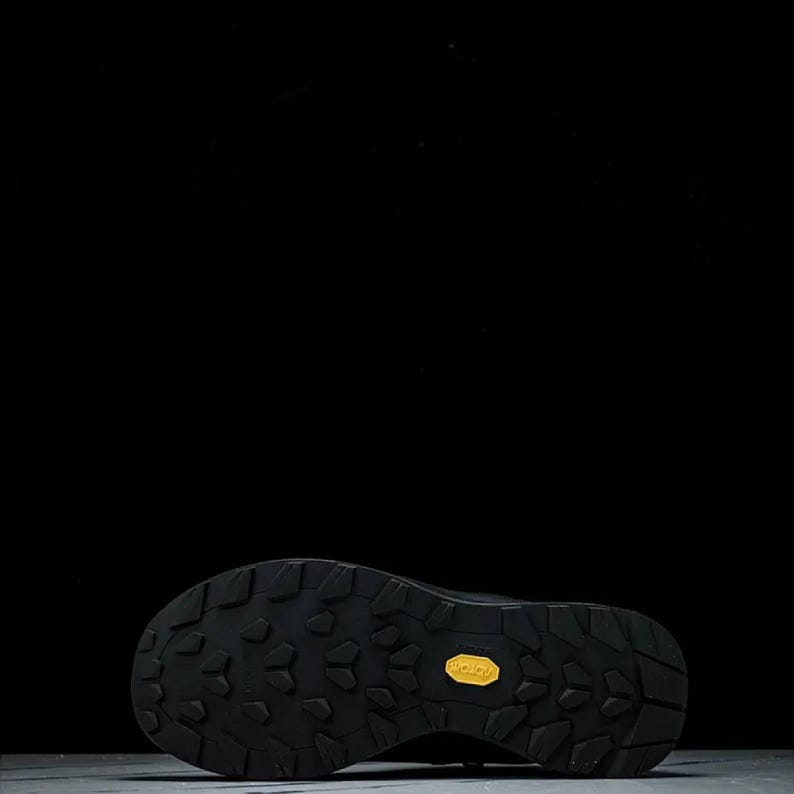 May include: Black hiking boot sole with a textured pattern for grip. The sole features a yellow Vibram logo. The image is set against a black background, highlighting the boot's design.