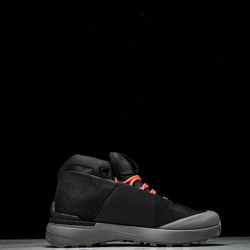 May include: Black and grey hiking boot with orange laces. The boot features a mesh upper and a grey sole. The design is modern and sleek, suitable for outdoor activities. The boot is shown against a black background.