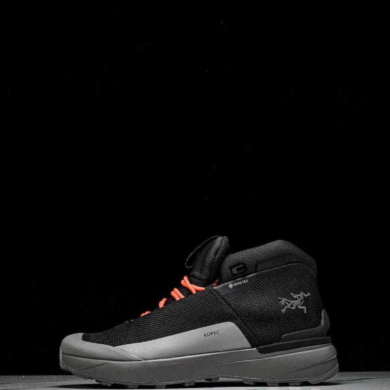 May include: Black and grey hiking boot with orange laces. The boot features a mesh upper, a grey sole, and the word "KOPEC" is printed on the side. The Arc'teryx logo is visible. Designed for outdoor activities, it has a Gore-Tex label.