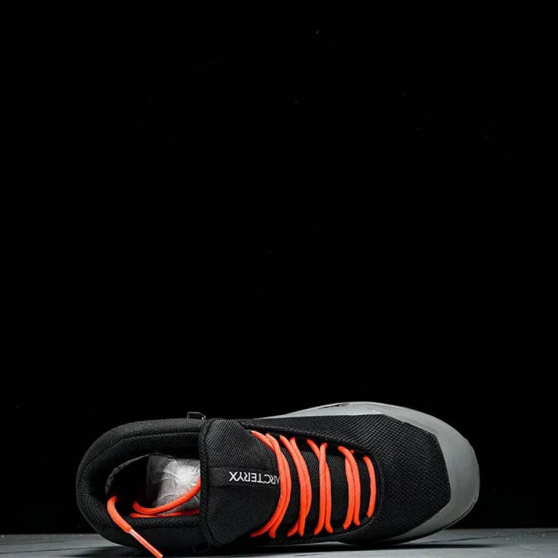 May include: Black and grey hiking boot with bright orange laces. The boot has a mesh upper and a grey toe cap. The brand name "Arc'teryx" is visible on the side. Designed for outdoor activities, it measures approximately 25 cm in length.