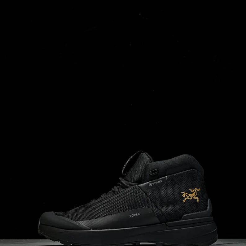 May include: Black high-top hiking boot with a textured upper and a thick sole. The boot features a gold logo on the side and the word "KOPEC" printed on the side. The boot is designed for outdoor activities.