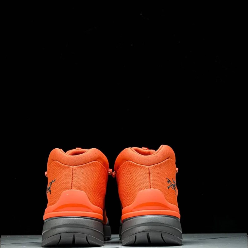 May include: Pair of vibrant orange hiking boots with dark grey soles, seen from behind. The boots feature a textured upper and a small logo on the side. The background is black.
