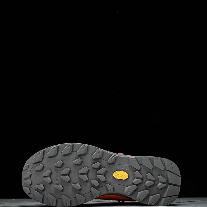 May include: Close-up of the sole of a hiking boot. The sole is grey with a textured pattern and a yellow "Vibram" logo. The boot's upper is a reddish-orange colour.