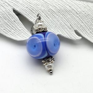 Könnte beinhalten: Ein blauer und weißer Glasperlenanhänger mit silberfarbenen Metallakzenten. Die Perle hat eine runde Form mit einem Wirbeldesign. Der Anhänger hat eine perlenartige Perle an der Unterseite und eine Schlaufe zum Befestigen.