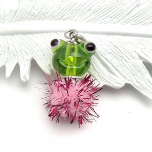 Könnte beinhalten: Ein skurriler Anhänger mit einer grünen Glasfroschfigur mit dunklen Augen und einem lächelnden Gesicht. Der Frosch ist an einem rosa, flauschigen Pompon mit metallisch roten Akzenten befestigt. Der Anhänger hängt an einer silberfarbenen Metallschlaufe.
