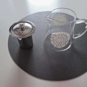 Könnte beinhalten: Eine transparente Teekanne aus Glas mit Griff und Ausguss, verziert mit einem weißen Blumenmuster. Ein Tee-Infuser aus Edelstahl mit Deckel steht neben der Teekanne auf einer dunkelgrauen runden Tischmatte.