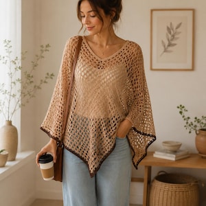 May include: A tan crochet poncho with a V-neck and dark brown trim. The poncho is worn over a light-colored top and paired with light blue jeans. A brown leather bag and a coffee cup are also visible.