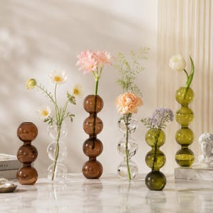 May include: A collection of decorative glass vases in various colors, including amber, clear, and olive green. Each vase features a unique stacked bubble design and holds fresh flowers, creating an elegant display. The vases are arranged on a marble surface.