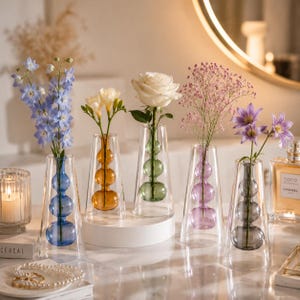 May include: Five glass vases with a unique design, each holding a different flower arrangement. The vases feature a clear, conical shape with three colored spheres stacked inside. The flowers include blue, white, and purple blooms.