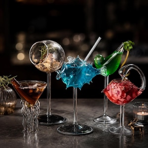 Può includere: Una collezione di bicchieri da cocktail unici, ognuno con un design distintivo. Un bicchiere ha la forma di un pesce, un altro di una sfera e altri hanno design di stelo creativi. I bicchieri sono riempiti con bevande e guarnizioni colorate.
