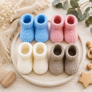 May include: Four pairs of handmade crochet baby booties arranged on a round tray. The booties are featured in solid colours: light blue, soft pink, cream white, and light brown. Each pair has a thick ribbed cuff and a textured knit pattern.