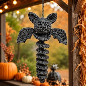 May include: A dark gray crocheted bat hanging ornament featuring a round head with two pointed ears, small black eyes, and two white fangs. The bat has outstretched wings and a long, coiled, spring-like body made of textured yarn stitches.