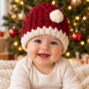 Pode incluir: Um gorro de crochê em vermelho escuro e branco creme com um padrão de malha texturizado e um único pompom cor de creme preso na lateral. O gorro apresenta uma aba larga e canelada na cor creme que contrasta com a coroa vermelha.