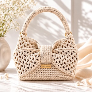 May include: A beige crochet handbag featuring a bow-shaped front design with an open-work lattice pattern and a solid base. The bag has a single rounded handle attached with gold-tone rings and a small rectangular metal tag labeled Mari Tejidos.