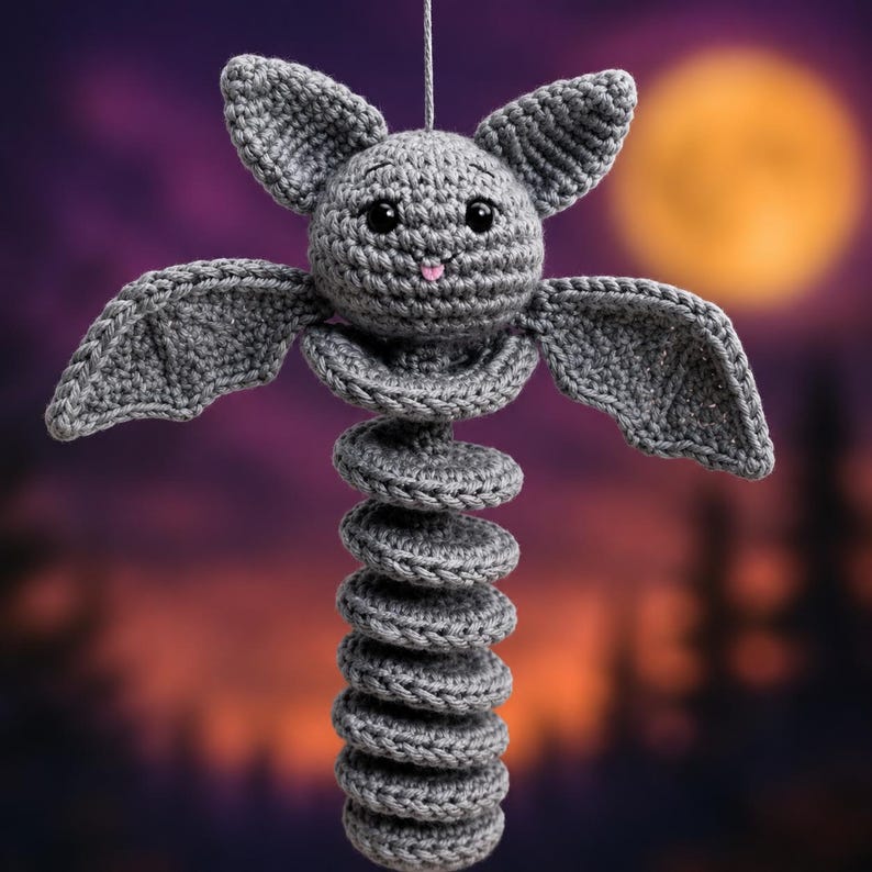 May include: A gray crocheted bat hanging ornament featuring a round head with pointed ears, large black safety eyes, and a small pink tongue. The body consists of a long, coiled, spring-like structure extending downward from the bat's wings.