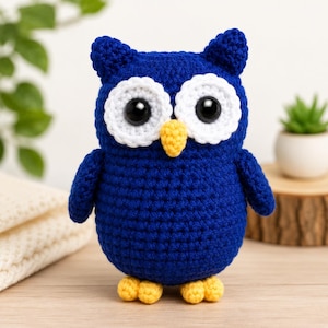 May include: A handcrafted crochet owl toy featuring a deep blue body, two large white circular eye patches with black plastic safety eyes, a small yellow beak, and two yellow feet. The owl has two small pointed ear tufts on top of its head.