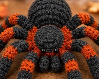 May include: A crocheted spider toy featuring a round black body and eight legs with alternating bands of black and burnt orange yarn. The spider has two small, smooth, black bead eyes positioned on the front of its head section.