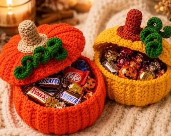 May include: Two crocheted pumpkin-shaped containers, one orange and one yellow, each featuring a stem and green curly vines. The orange container has a lid and is filled with assorted candy bars like Twix, Snickers, and KitKat. The yellow container is open and contains various wrapped candies.