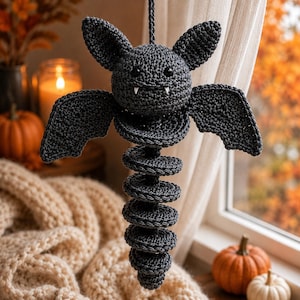 Può includere: Un pipistrello decorativo realizzato all'uncinetto in grigio scuro, con testa rotonda, due orecchie a punta, occhi a perla neri e due piccole zanne bianche. Il corpo è formato da una lunga coda segmentata a spirale che pende verticalmente.