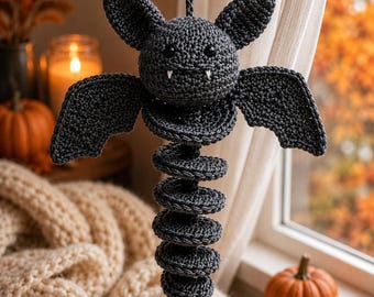 May include: A dark gray crocheted bat ornament featuring a round head with two pointed ears, black bead eyes, and two small white fangs. The body consists of a long, spiraling, segmented tail hanging vertically from the center of the wings.