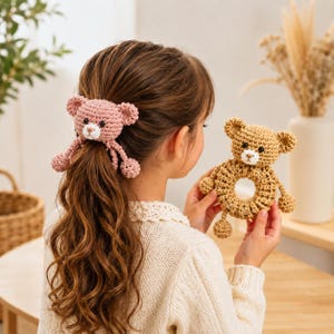 Puede incluir: Una goma de pelo de oso de ganchillo en color rosa empolvado con orejas pequeñas y hocico blanco, junto a un sonajero de anilla de oso de ganchillo en color tostado con centro circular, ojos oscuros y hocico blanco.