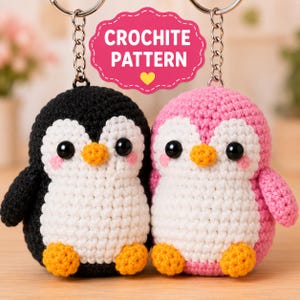 May include: Two crochet penguin keychains with metal rings. One penguin is black and white, the other is pink and white. Both have orange beaks and feet, black eyes, and pink cheeks. A pink sign above them reads CROCHITE PATTERN with a yellow heart.