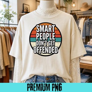 Op de afbeelding: Crèmekleurig T-shirt met een cirkelvormige afbeelding met de tekst "SMART PEOPLE DON'T GET OFFENDED" in witte letters. De afbeelding heeft een retro-ontwerp met oranje, rode en turquoise strepen. Het T-shirt wordt op een mannequin getoond.