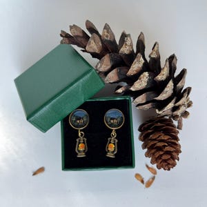 Peut inclure: Une paire de boucles d'oreilles pendantes avec un haut circulaire représentant un paysage nocturne peint d'une maison et une breloque en forme de lanterne ancienne. Les boucles sont présentées dans une boîte verte carrée à intérieur velours noir.