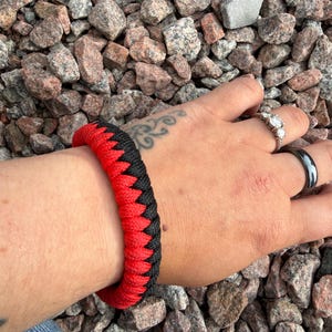 May include: A red and black braided bracelet. The bracelet is made of a thick, woven material, with a zig-zag pattern where the red and black colours meet. The bracelet is shown on a wrist, against a background of small rocks.