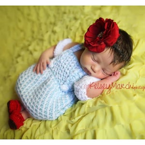 Puede incluir: Un bebé con un vestido de crochet azul y blanco con una diadema de flores rojas, durmiendo en una manta amarilla con volantes.