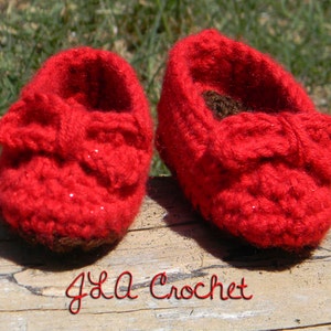 Op de afbeelding: Een paar rode gehaakte babyschoentjes met een strik op elke schoen. De schoentjes zijn gemaakt van een glinsterend rood garen.