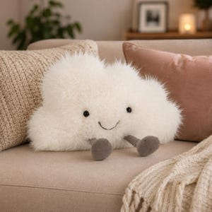 May include: A soft, white cloud-shaped plush toy with black button eyes and a stitched smile. It has grey, rounded feet. The plush is on a beige sofa with decorative pillows and a blanket.
