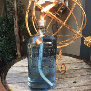 May include: A clear glass bottle with a blue liquid inside. The bottle has a label that reads "BULLEIT BOURBON" and is being used as a decorative torch. The bottle is sitting on a wooden surface.