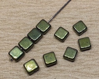 May include: A group of small, square, metallic beads in a green and gold color. The beads are scattered on a brown surface, with some of them strung on a thin wire.