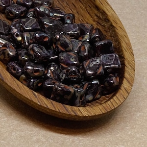 Op de afbeelding: Een houten kom gevuld met donkere, glanzende kralen. De kralen zijn een mix van zwart, paars en oranje, met een gespikkeld patroon. De kom is ovaal van vorm en gemaakt van donker hout met zichtbare nerf.