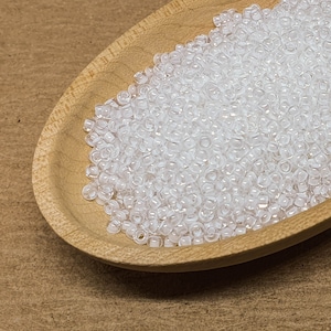 May include: A wooden bowl filled with clear glass seed beads. The beads are small and round, and they are arranged in a random pattern.