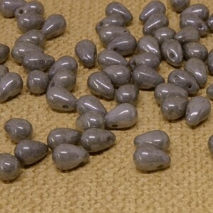 Könnte beinhalten: Eine Sammlung grauer, tropfenförmiger Glasperlen. Die Perlen sind auf einer beigefarbenen Stoffoberfläche verstreut.