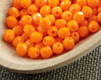 Puede incluir: Una cuchara de madera llena de cuentas de color naranja brillante. Las cuentas son pequeñas, redondas y tienen un acabado brillante. La cuchara es de madera clara y está colocada sobre un fondo gris texturizado. Las cuentas son probablemente para manualidades o joyería.