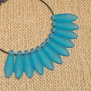 May include: A necklace with a dark metal hoop and a series of light blue, teardrop-shaped beads. The beads are arranged in a fan-like pattern, creating a unique design. The necklace is set against a neutral, textured background.