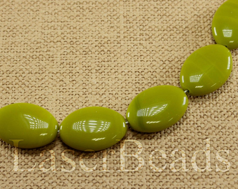 May include: A strand of oval-shaped green beads on a brown fabric background. The beads are smooth and glossy.