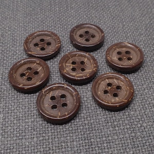 Könnte beinhalten: Sieben braune Kokosnuss-Knöpfe mit jeweils vier Löchern. Die Knöpfe sind in einem Halbkreis auf einem braunen, strukturierten Stoff angeordnet.