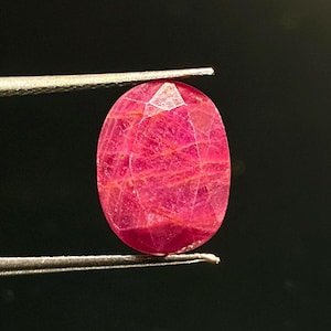 Puede incluir: Una piedra preciosa de rubí de corte ovalado con un tono rojo rosado intenso e inclusiones internas visibles, sostenida por unas pinzas de metal plateado sobre un fondo oscuro y sólido. La piedra presenta una superficie facetada que refleja luz.
