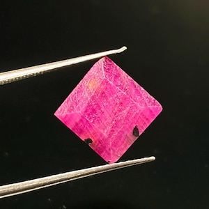 May include: A square-cut, vibrant pink gemstone held by silver-toned metal tweezers. The stone features a faceted surface with visible internal textures and two small, distinct black inclusions near the bottom corners of the square shape.