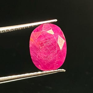 以下が含まれることがあります： 鮮やかなピンクがかった赤色の色合いと多面的な表面を持つオーバルカットのルビーの宝石。銀色の金属製ピンセットの先端でしっかりと保持されており、暗い背景に対してその内部の透明度と質感が際立っています。